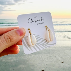May include: Cream-colored seashell earrings with gold-colored hardware. The earrings are displayed on a white card with the text "727 Clayworks JEWELRY". The background shows a beach scene with the ocean and a sunset.