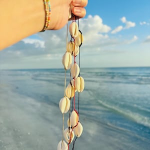 Puede incluir: Una colección de collares de conchas blancas colgando contra un fondo de cielo azul y océano. Los collares están ensartados en cordones negros, rojos y beige. Una pulsera de cuentas coloridas es visible en la esquina superior izquierda.