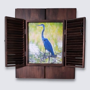 May include: A wooden shutter frame with a blue heron standing in a marsh, the shutters are open and the image is framed in a brown wood frame.