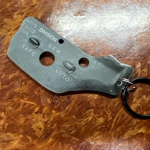 May include: A silver metal keychain with a black ring. The keychain has the words "ARMS DIVISION" and "SAFE, SEMI, AUTO" engraved on it.