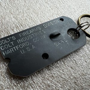 May include: A black metal key chain with the text "Colt's Firearms Division Colt Industries Hartford, Conn. U.S.A." and "Safe" printed on it. The key chain has a brass ring and a small hole for a key.