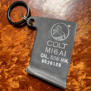 May include: A black metal keychain with a Colt M16A1 logo, a running horse, and the text "CAL. 5.56 MM. 9536129".