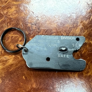 May include: A silver metal key chain with a black ring. The keychain has the text "Colt Industries Division S. Firearms Hartford, Conn. U.S.A." and "Safe" on it.
