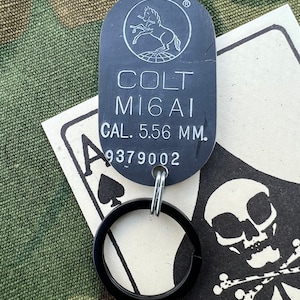 May include: Black metal keychain with a Colt M16A1  5.56 mm.  tag and a black ring. The tag has a Colt logo with a rearing horse and the text "Colt M16A1 CAL. 5.56 MM. 9379002".