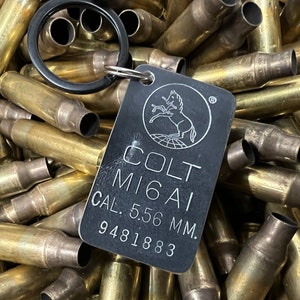 May include: A black metal keychain with a Colt M16A1 5.56 mm caliber logo and a silver ring. The keychain is surrounded by a pile of brass casings.