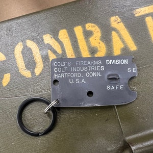 May include: A black metal keychain with a silver ring. The keychain has the text "Colt's Firearms Division Colt Industries Hartford, Conn. U.S.A. Safe" on it.