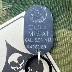 May include: A black metal keychain with a Colt M16A1  5.56 mm  dog tag and a serial number 9498129. The dog tag has a Colt logo with a rearing horse and a circle around it.