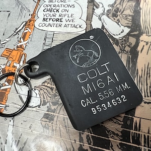 May include: Black metal keychain with a Colt MI6AI inscription, a silver horse logo, and the text "CAL. 5.56 MM. 9534632".