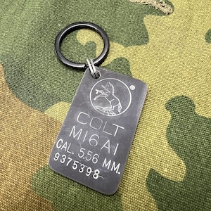 May include: A silver metal keychain with a black ring. The keychain has the text "COLT M16A1 CAL. 5.56 MM. 9375398" engraved on it.