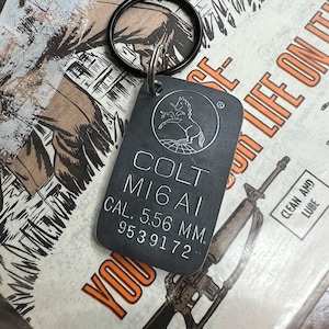 May include: A black metal keychain with a silver tag that reads "COLT MIGAI CAL. 5.56 MM. 9539172". The tag features a stylized image of a horse and a globe.