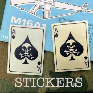 May include: Two black and white stickers of the Ace of Spades playing card with a skull and crossbones design. The stickers are on a camouflage background.