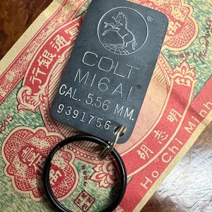 May include: A black metal keychain with a silver Colt M16A1 logo and the text "CAL. 5.56 MM." and the serial number "9391756".