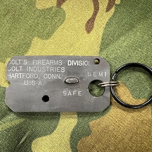 May include: A rectangular, gray metal tag with the text "COLT'S FIREARMS DIVISION, COLT INDUSTRIES, HARTFORD, CONN. U.S.A." and "SAFE" printed on it. A black key ring is attached. The background is a camouflage pattern.