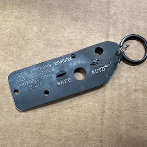 May include: Black metal key fob with the text "Colt's Firearms Division Colt Industries Hartford, Conn. U.S.A." and the words "Safe", "Semi", and "Auto" printed on it. The key fob has a black metal ring attached.