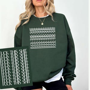 May include: Dark green sweatshirt with a white graphic design and a white crew neck. The graphic design is a series of horizontal lines and shapes. A card with the text "FUCK THIS SHIT" is in the image.