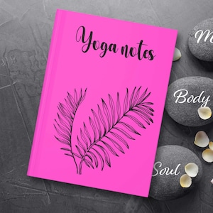 Könnte beinhalten: Rosafarbenes Yoga-Notizbuch mit einer schwarzen Palmenblatt-Illustration. Das Notizbuch ist von grauen Steinen umgeben, auf denen die Wörter "Mind", "Body" und "Soul" stehen.