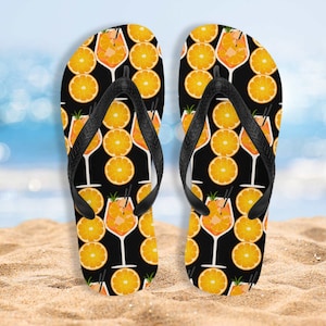 May include: Pair of black flip flops with a pattern of orange slices and cocktails on a black background.