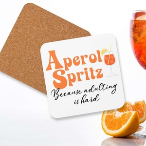 Könnte beinhalten: Weißer Untersetzer mit dem Text "Aperol Spritz Because adulting is hard" in schwarzer und orangefarbener Schrift. Neben dem Text ist ein Glas Aperol Spritz mit Eis und einer Orangenscheibe abgebildet.