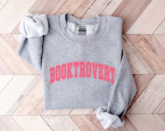May include: A grey sweatshirt featuring the word "BOOKTROVERT" in bright pink lettering. The garment is folded on a light brown wooden surface. The sleeves are rolled up.