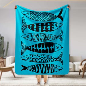 May include: A turquoise throw blanket featuring a repeating pattern of stylized black fish. Each fish has a unique design, with details like stripes, dots, and geometric shapes. The blanket is held up, showcasing its full design.