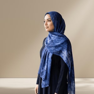 May include: A woman wearing a dark blue hijab and a matching scarf. The scarf has a blue and white marbled pattern. The woman is wearing a black long-sleeved top. The background is a solid beige color.
