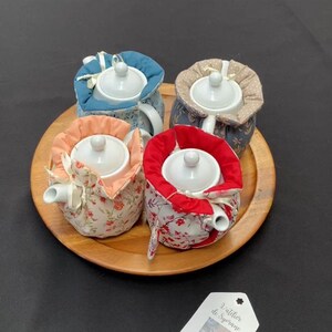 Peut inclure: Quatre théières en céramique blanche, chacune enveloppée dans une housse en tissu avec des motifs distincts et des doublures unies bleu, beige, pêche et rouge. Les théières sont disposées sur un plateau rond en bois avec une étiquette blanche.
