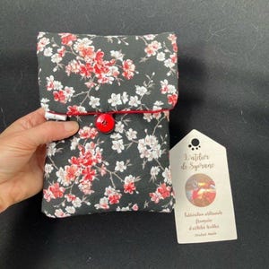 Pode incluir: Uma bolsa de tecido retangular com fundo preto e um padrão floral de flores de cerejeira brancas e vermelhas. A bolsa tem uma aba dobrável presa por um botão vermelho. Uma etiqueta branca com o texto L'atelier de Syerane está anexada.