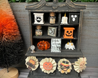May include: A black Halloween-themed display shelf with various miniature decorations. The shelf contains small boxes with faces, bottles, a pumpkin, and a banner with Halloween characters. A small orange and black tree is on the left.