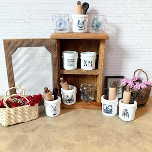 May include: A miniature kitchen scene with a wooden shelf and various decorative items. There are small white ceramic jars with blue illustrations, a wire basket with red roses, and a small framed picture. The scene evokes a rustic, vintage aesthetic.