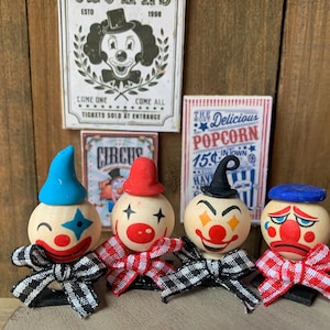 May include: Four wooden clown figurines with blue, red, and black hats. Each clown has a different facial expression and is wearing a red and white checkered bow tie. The clowns are standing on a wooden surface in front of a wall with vintage circus posters.