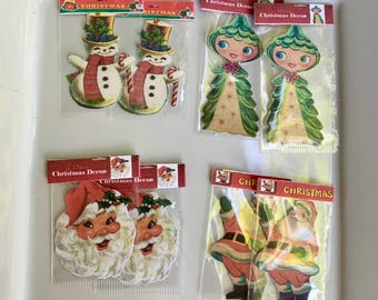 May include: Four sets of vintage-style Christmas paper cutouts in clear plastic packaging. The designs include snowmen with top hats, green tree-shaped figures, Santa Claus faces, and full-body Santa figures, all labeled with Christmas Decor text.