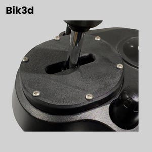 Könnte beinhalten: Ein schwarzer Kunststoff-Schalthebelsockel mit einem silbernen Schalthebel und einer runden schwarzen Platte mit mehreren Schrauben. Die Platte hat einen Schlitz für den Schalthebel.