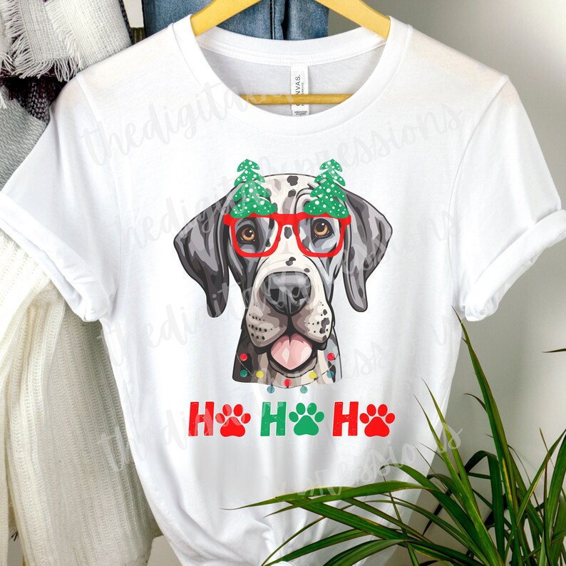 May include: White t-shirt with a cartoon illustration of a Great Dane wearing red glasses with Christmas trees on top and a string of Christmas lights around its neck. The text "Ho Ho Ho" is written below the dog with paw prints replacing the letter "O".