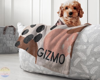 Op de afbeelding: Een kleine hond met krullend haar kijkt over een dierenbed. Het bed heeft een deken met abstracte vormen in bruin, zwart en perzik, met de naam "GIZMO". Het bed is grijs met een veermotief.