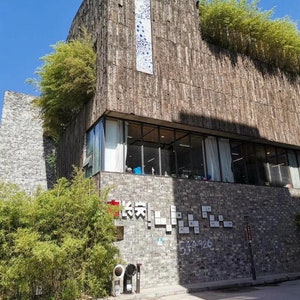 May include: A multi-story building with a textured, brown facade and a flat roof with green plants. The building has large windows and a sign with Chinese characters. A blue sky is visible in the background.