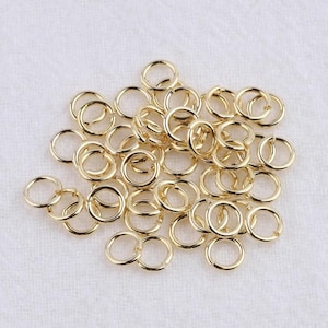 May include: A collection of small, gold-colored jump rings scattered on a white surface. The rings are circular and appear to be made of metal, with a shiny finish. They are likely used for jewelry making or other craft projects.