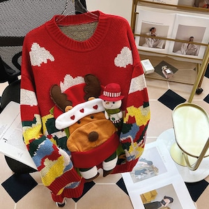 Könnte beinhalten: Roter Weihnachtspullover mit einem weißen und blauen Rentier- und Schneemann-Design. Der Pullover hat einen Rundhalsausschnitt und lange Ärmel.