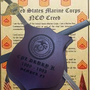 May include: A framed display featuring a US Marine Corps NCO Creed document, a sword, and a wooden plaque. The plaque is engraved with the Marine Corps emblem, the name "CPL DRUKP H.", and the years 1989-1993. The sword has a gold hilt.