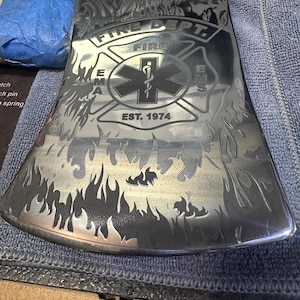 May include: A polished metal fire axe with engraved details. The axe head features the text "FIRE DEPT." and "EMS" with a medical symbol and the year "EST. 1974." Flames are etched along the blade's edge, creating a striking design.