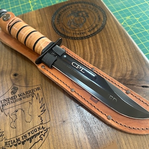 May include: A black combat knife with a leather sheath, resting on a wooden plaque. The knife has the initials "CPL" etched on the blade. The plaque features the US Marine Corps emblem and text.