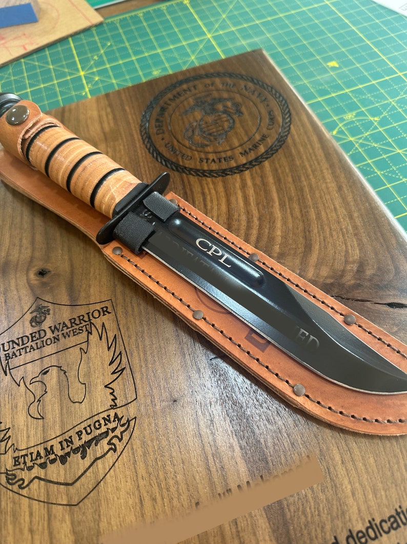 May include: A black combat knife with a leather sheath, resting on a wooden plaque. The knife has the initials "CPL" etched on the blade. The plaque features the US Marine Corps emblem and text.
