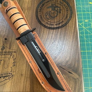 May include: A black combat knife with a leather handle and sheath, resting on a wooden plaque. The plaque features the US Marine Corps emblem and text. The knife blade is inscribed with "CPL".