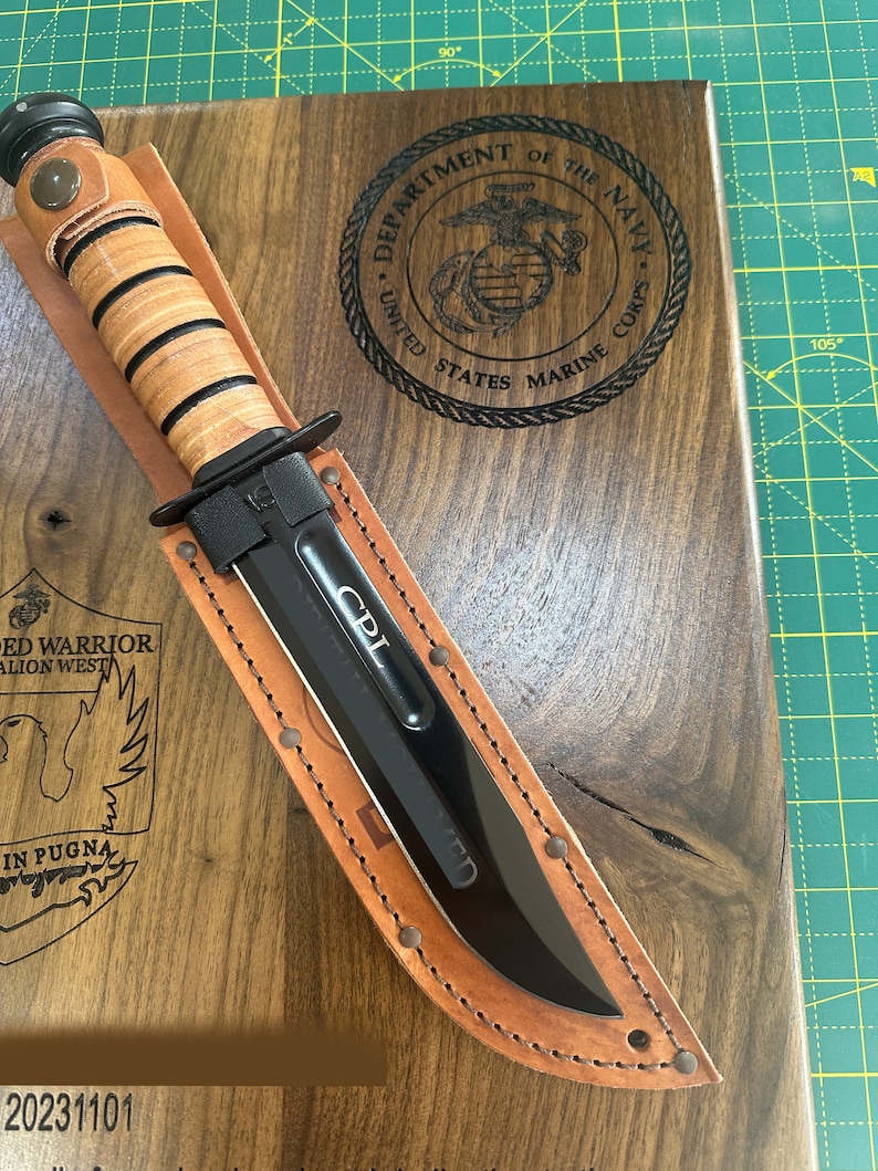 May include: A black combat knife with a leather handle and sheath, resting on a wooden plaque. The plaque features the US Marine Corps emblem and text. The knife blade is inscribed with "CPL".