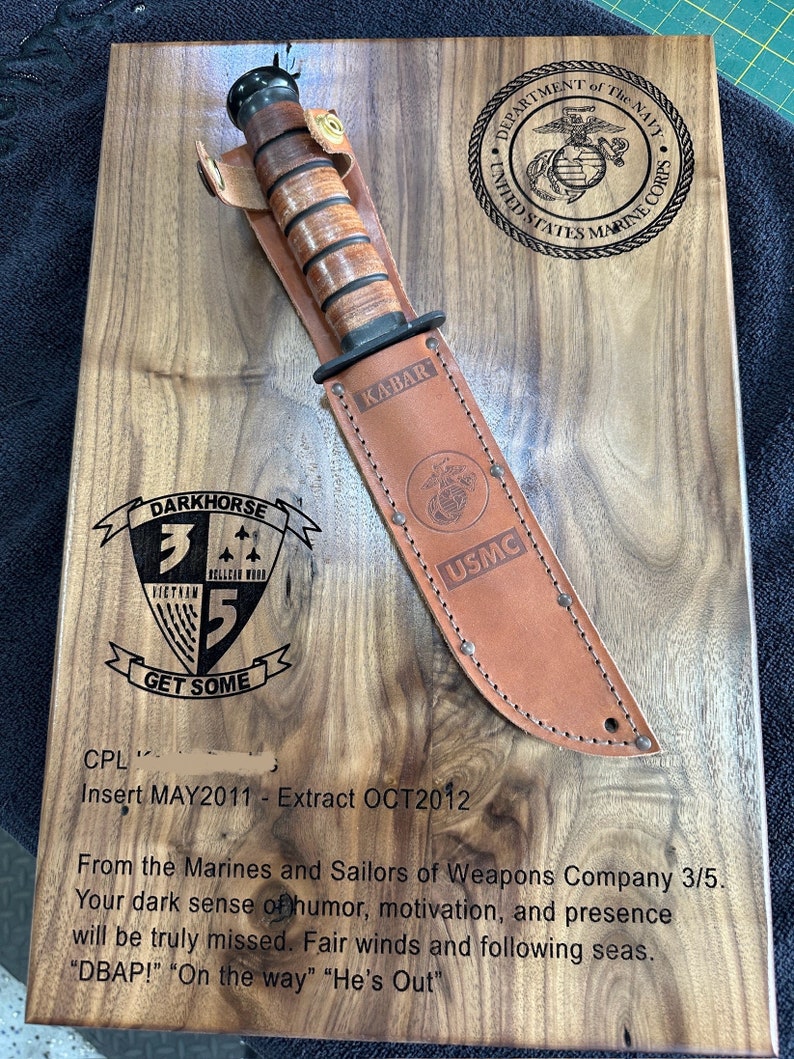 May include: A wooden plaque with a Ka-Bar knife in a leather sheath. The plaque has engraved text and emblems, including "DEPARTMENT of The NAVY UNITED STATES MARINE CORPS" and "DARKHORSE 3/5 GET SOME". The knife has a brown handle and a leather sheath with "USMC" printed on it.