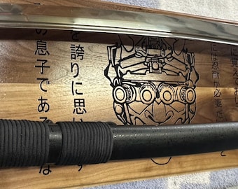 May include: A black katana sword with a black and grey handle, resting on a wooden display stand. The stand features a laser-engraved design of a samurai warrior's face with a helmet and goggles, and Japanese text that reads "に覚えておいてくださいには忍耐が必要だ誇りに思いの息子であっは".
