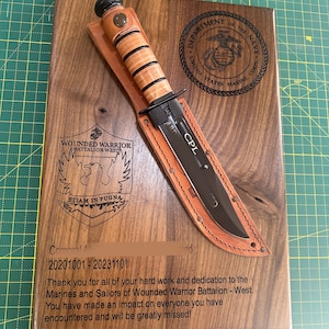 May include: A wooden plaque with a knife and sheath. The plaque has engraved text and the US Marine Corps emblem. The knife has a black blade and a tan handle. The text includes "Wounded Warrior Battalion West" and "CPL".