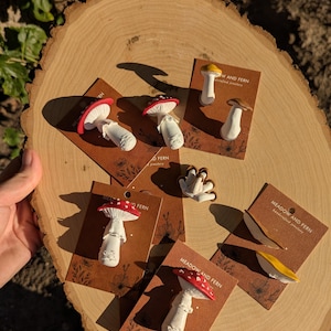 May include: A collection of mushroom-shaped jewelry pieces, including pins and earrings, displayed on small brown cards. The mushrooms feature red, white, and yellow caps with detailed stems. The cards are arranged on a wooden slice, with the text "MEADOW AND FERN" visible.