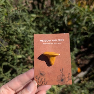 May include: A mustard-yellow mushroom pin on a brown card with the text "MEADOW AND FERN handcrafted jewelery". The card has floral illustrations. The pin is made of a light-coloured material.