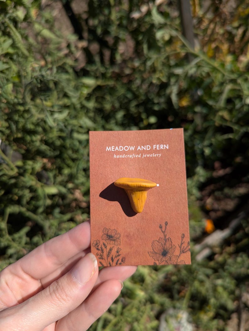 May include: A mustard-yellow mushroom pin on a brown card with the text "MEADOW AND FERN handcrafted jewelery". The card has floral illustrations. The pin is made of a light-coloured material.