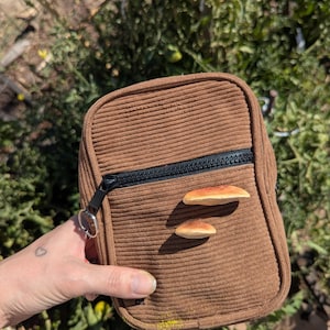 May include: A brown corduroy crossbody bag with a black zip and two mushroom decorations. The bag is rectangular with a small silver keyring attached to the zip. The background is blurred green foliage.